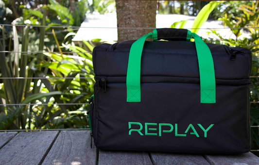 A black bag with the word REPLAY on it in green sits on a wooden bench in front of some plants