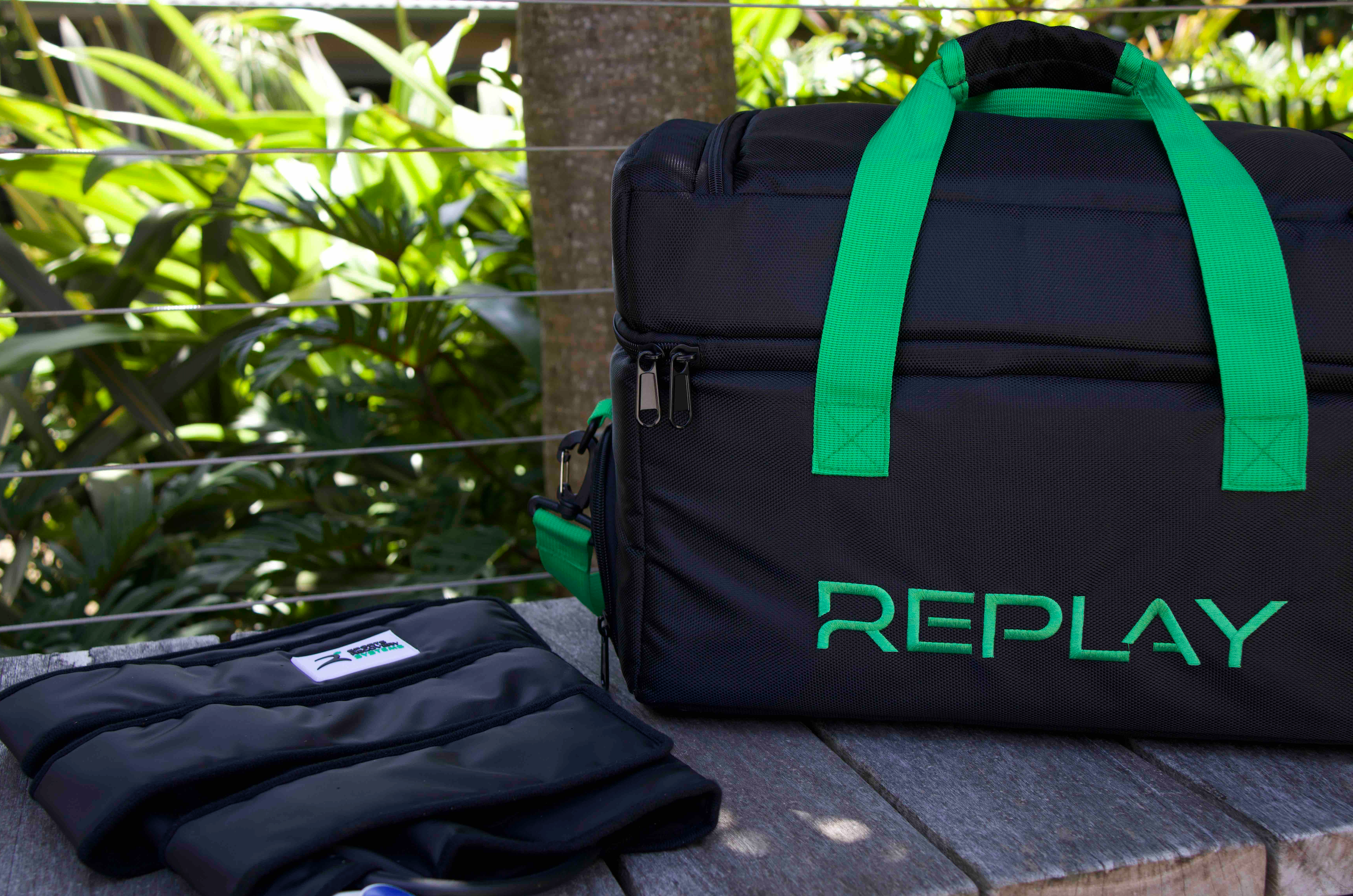 A black bag with the word Replay in green and knee compression pad sit on a wooden bench with plants in the background