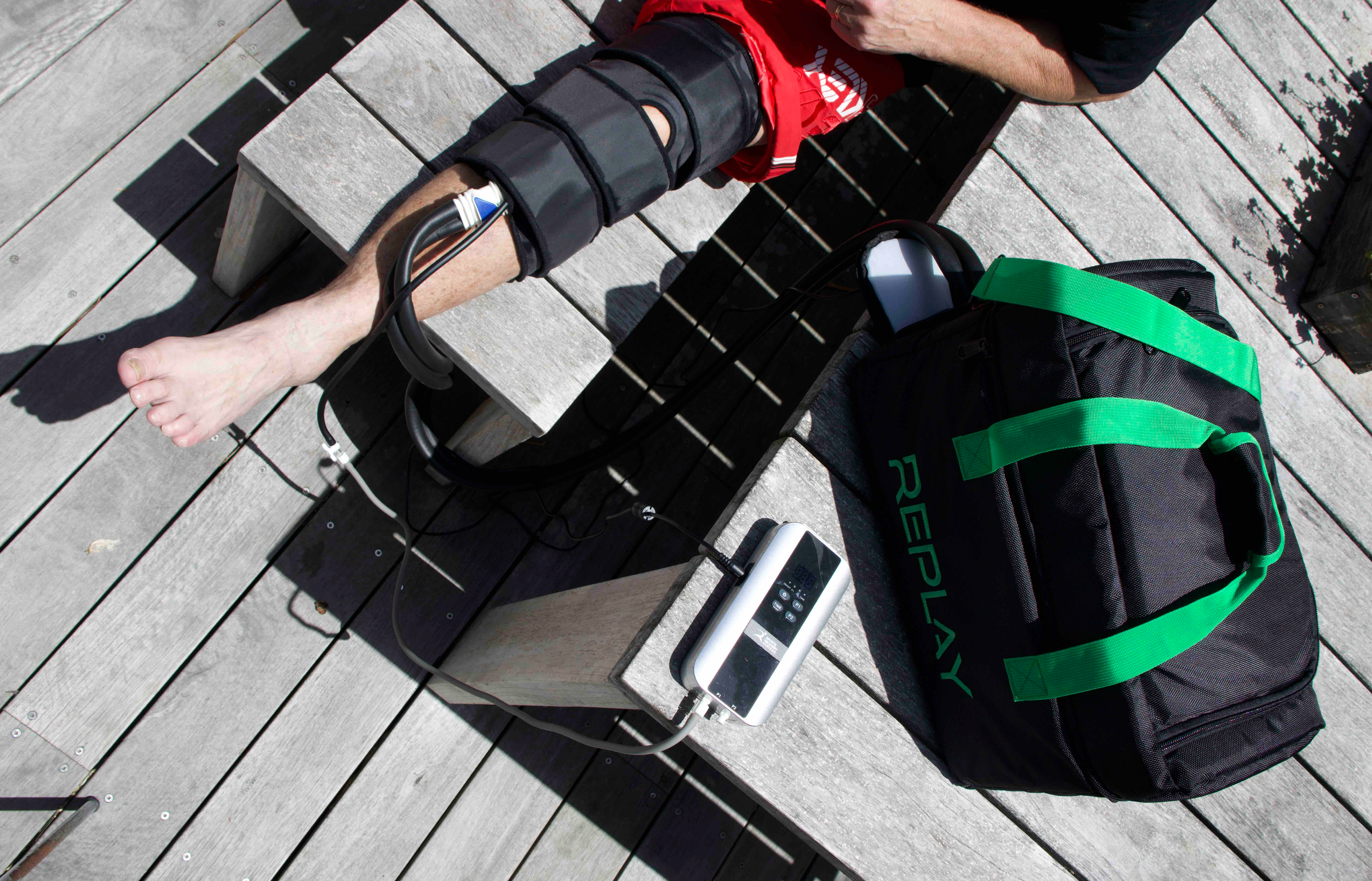A man's leg is seen with ice compression therapy equipment on knee with black bag next to showing the word "Replay" in green