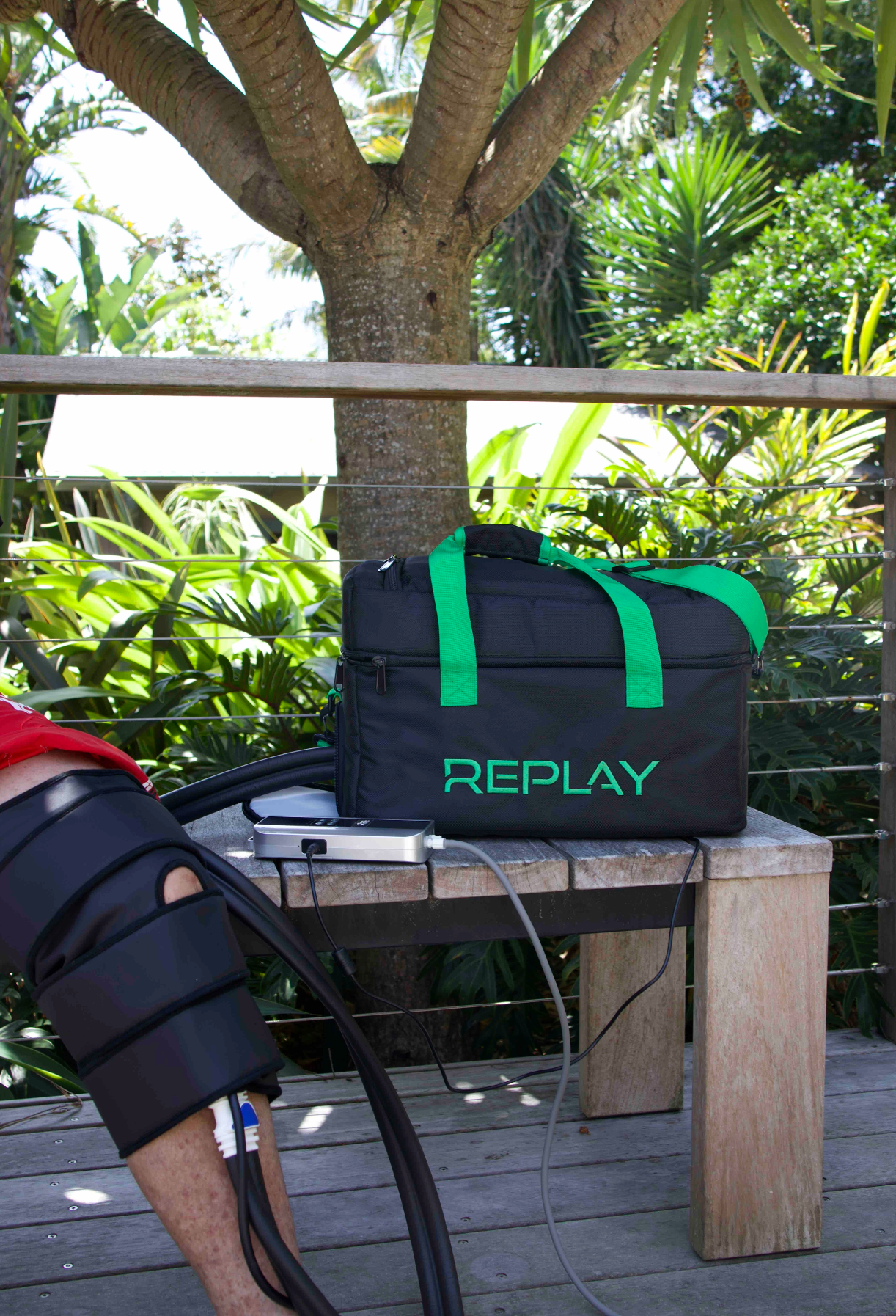 A man's knee with compression pad is shown next to a black bag with the word Replay in green sitting on a wooden bench with plants in the background
